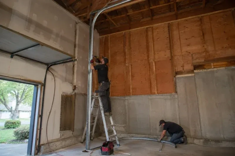 One person on a stepladder working beside a tall curved garage-door track while another kneels beside loose track sections in an unfinished garage. — High-Lift Garage Door Conversion, East Gwillimbury